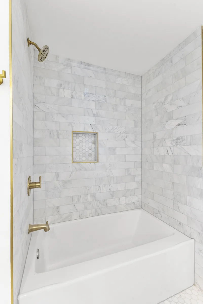 Carrara tub surround. Running bond to the ceiling, brass edge profile, hex mosaic set into the niche.